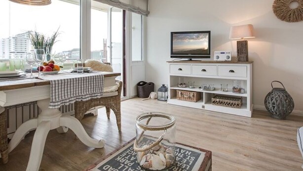 A modern living room with a dining area, large windows, a television, and a white cabinet with shelves. - New Port II - Newport on the beach with south-facing balcony and dune view (Sylt)