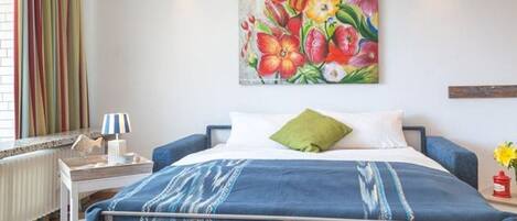 A bedroom with a metal bed frame, a blue blanket, a green pillow, and a floral painting on the wall.