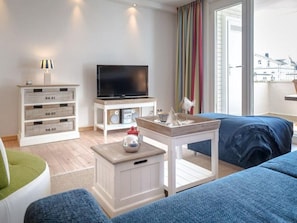 A modern living room with a flat-screen TV, a coffee table, and a sofa. There are storage units with labeled drawers and a window with curtains. - Blue Water - House Eidum, on the beach, 2 room apartment with balcony (Sylt)