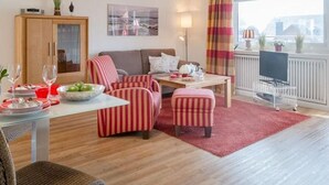 A modern living room with a sofa, striped armchairs, a dining table with plates and glasses, and a television on a stand. - Nordstern - 1-room apartment with balcony and beach chair in the immediate vicinity (Sylt)