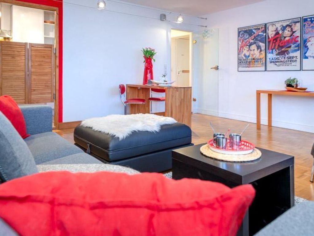 Una sala moderna con un sofá, un reposapiés y una mesa de centro. Hay una zona de comedor con una mesa y sillas. La habitación tiene una pared de acento roja y un cartel de una película.
