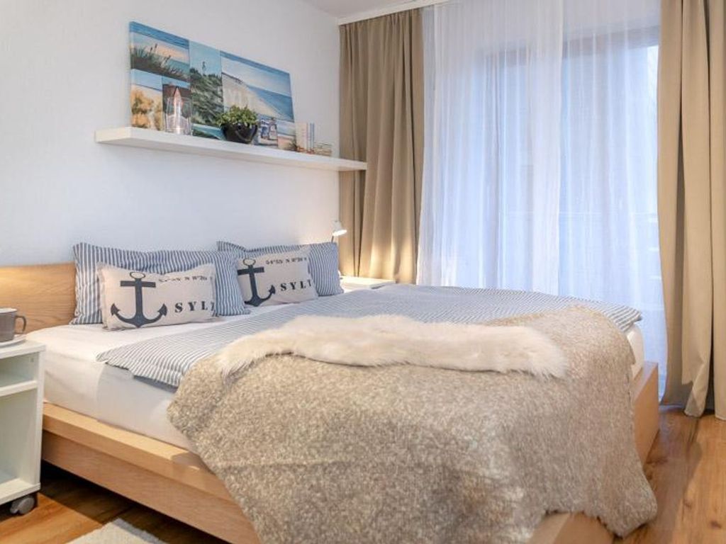 A bedroom with a bed, a nightstand, a shelf with decorative items, and a window with curtains.