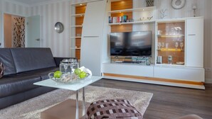 Un salon moderne comprenant un canapé, une table basse en verre agrémentée d’objets décoratifs, un téléviseur à écran plat et des étagères intégrées où sont disposés des livres et d’autres objets décoratifs.