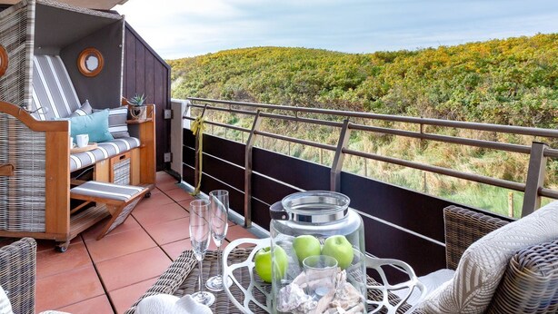 Un balcon agrémenté de meubles en osier, une table avec des verres et des pommes, et une vue panoramique sur une colline boisée.