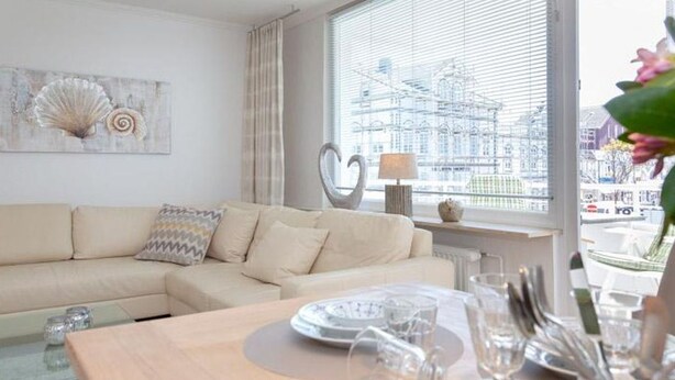 En stue med en beige sofa, et spisebord dækket med tallerkener, glas og bestik, samt et stort vindue med udsigt til bygninger.