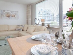 A living room with a beige sofa, a dining table set with plates, glasses, and cutlery, and a large window with a view of buildings. - Atlantik 40 - High-quality furnished, 3-room apartment in a prime location. (Sylt)