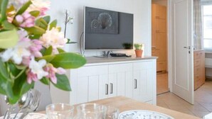 Et spiseområde med et bord dækket til fire personer, et fladskærms-TV monteret over disken, og en potteplante på disken.