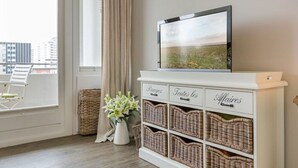 A room with a white console table featuring wicker baskets labeled "Rameaux," "Toutes les," and "Affaires." A flat-screen TV is mounted above the table, and a window with a view of buildings is visible.
