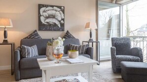 A modern living room with a grey sofa, a white coffee table, and a patterned armchair. There is a painting on the wall and a window with a view of buildings. - Viktoria Residenz 9 - Top city location: Very comfortable, light-flooded 2 (Sylt)