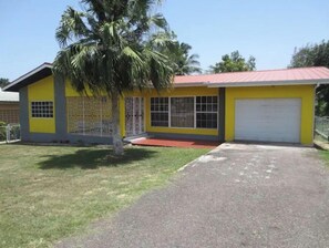 A single-story yellow building with a red roof, a garage, and a small front yard with a palm tree. - Chateau Yellow on Wint Close (Mandeville)