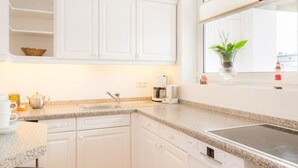 A modern kitchen with white cabinets, a granite countertop, a built-in dishwasher, and a window with a plant. - Kurt`s Island - In Friedrichstraße: 2.5 room apartment with balcony (Sylt)
