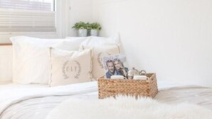 Ein ordentlich bezogenes Bett mit weißer Bettwäsche, ein flauschiger weißer Teppich und ein Korbwäschetablett mit Foto und Kleinigkeiten.