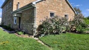 A stone building with a gabled roof, a satellite dish, and a small garden with flowering plants. - Primitive yet classy apartment in rural Iowa near small town shopping and hiking (Colesburg)