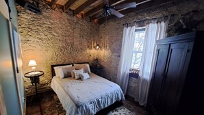 A bedroom with a stone wall, a bed with a white bedspread, a nightstand with a lamp, and a wooden ceiling fan. - Primitive yet classy apartment in rural Iowa near small town shopping and hiking (Colesburg)