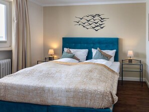 A bedroom with a blue headboard, two bedside tables with lamps, a patterned bedspread, and a wall art piece. - Dwarslöper 2 - 2 room apartment on the beach - Outstanding sea view from the 5th floor (Sylt)