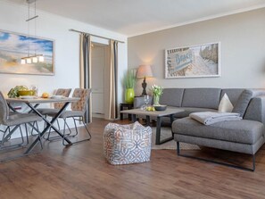 A modern living room with a grey sofa, a dining table with chairs, a coffee table, and a painting on the wall. - Dwarslöper 2 - 2 room apartment on the beach - Outstanding sea view from the 5th floor (Sylt)