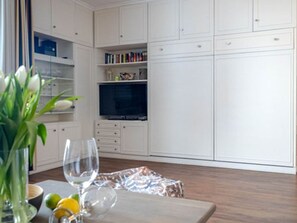 A room with a wooden floor, white cabinets, a television, and a table with a vase and glasses. - Dwarslöper 2 - 2 room apartment on the beach - Outstanding sea view from the 5th floor (Sylt)