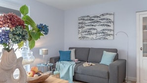 Una sala de estar con un sofá gris, una mesita de madera con un jarrón de flores, una obra de arte en la pared y una lámpara de pie.