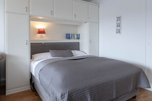 Een slaapkamer met een bed, nachtkastje, lamp en een plank met boeken.