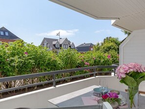 Un balcón con una mesa para dos, un florero con flores y vistas a casas y follaje.