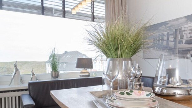 A dining area with a table set for two, a potted plant, and a view of the outside. - Beachwalk - Brandenburg House: 3-room apartment, 2 bedrooms, balcony, ideal for (Sylt)