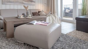 A modern living room with a sofa, ottoman, and coffee table, a large window with curtains, and a view of the outdoors. - Beachwalk - Brandenburg House: 3-room apartment, 2 bedrooms, balcony, ideal for (Sylt)
