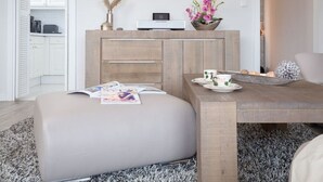 A modern living room with a sofa, ottoman, coffee table, and a dining area with a table and chairs. There is a kitchen area with cabinets and a microwave. - Beachwalk - Brandenburg House: 3-room apartment, 2 bedrooms, balcony, ideal for (Sylt)