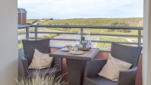 A balcony with wicker chairs and a table, overlooking a grassy landscape and a building in the distance. - Beachwalk - Brandenburg House: 3-room apartment, 2 bedrooms, balcony, ideal for (Sylt)