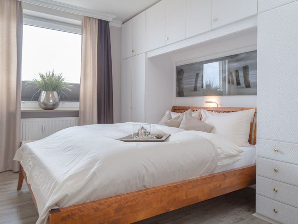 A bedroom with a wooden bed, white bedding, a bedside table with a tray, a window with curtains, and a wall-mounted artwork.