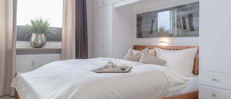 A bedroom with a wooden bed, white bedding, a bedside table with a tray, a window with curtains, and a wall-mounted artwork.