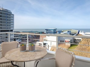 Un balcon avec une table ronde et deux plantes en pot, d’où l’on peut admirer un paysage urbain côtier avec ses bâtiments et la mer.