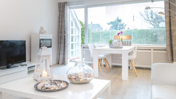 Una sala moderna con televisor de pantalla plana, una mesa de centro blanca con adornos, un comedor con mesa y sillas blancas, y un ventanal con vistas a zonas verdes y edificios.