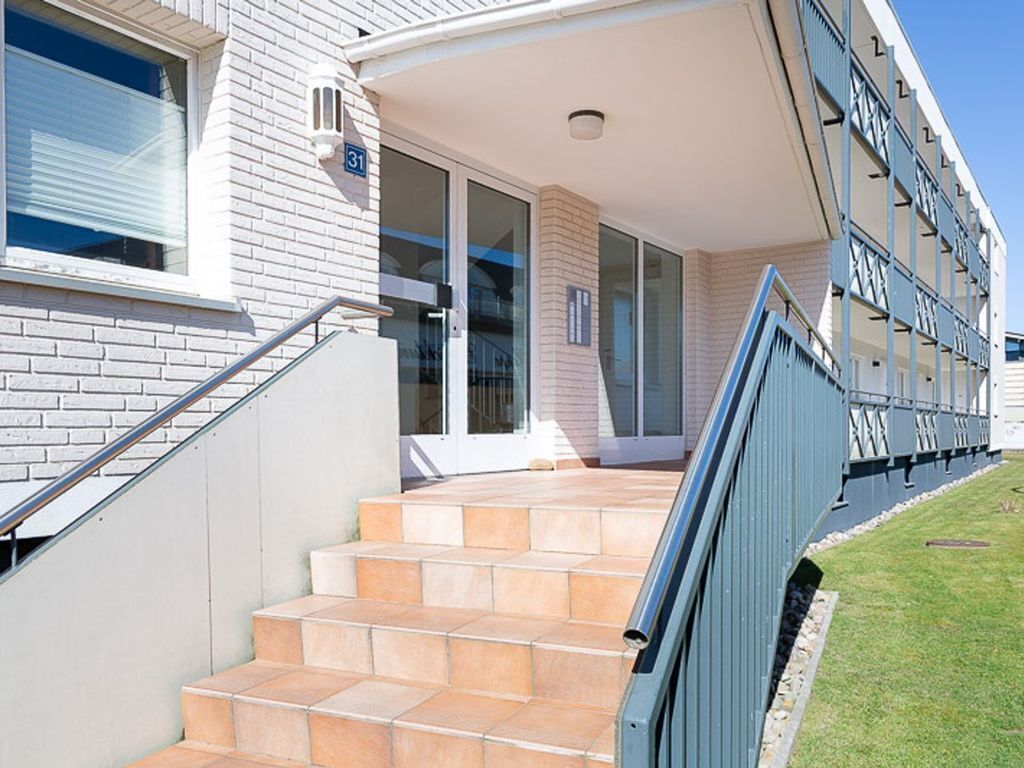 Un bâtiment moderne avec une façade en briques, un escalier recouvert de carreaux beiges menant à une entrée avec une porte en verre, et une rambarde bleue le long du côté.