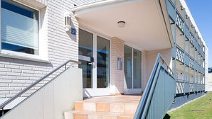 Ein modernes Gebäude mit einer Ziegel-Fassade, eine Treppe mit beigen Fliesen, die zu einem Glastür-Eingang führt, und ein blauer Geländer entlang der Seite.