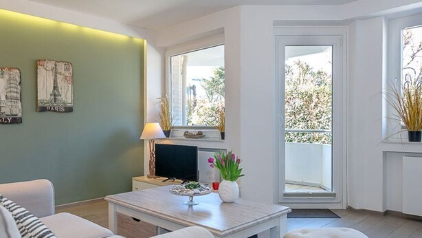 Ein modernes Wohnzimmer mit einer Couch, einem Hocker und einem Esstisch mit einer Lampe. Zwei Fenster bieten Blick auf Grünflächen und einen Balkon.