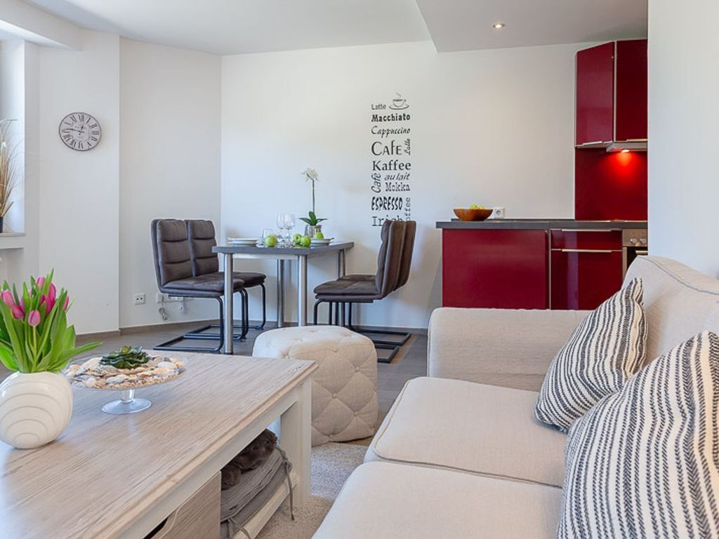 Un salon moderne avec un canapé, un coin repas avec des chaises, une petite table ornée d’un vase de fleurs et un coin cuisine avec des meubles rouges.