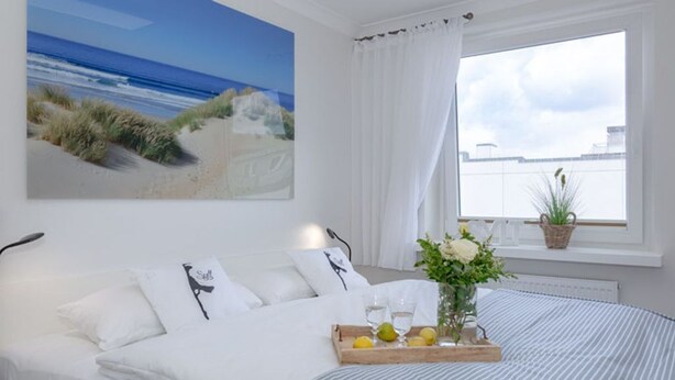 A neatly made bed with a striped bedspread, a bedside table with a tray of lemons and a vase of flowers, and a large window with a view of the beach and city skyline. - Black Pearl - Elisabethstraße, Dünenburg House, modern 2-room apartment with (Sylt)