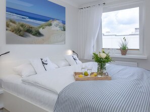 Een opgemaakt bed met een gestreept beddegoed, een nachtkastje met een schaal citroenen en een bloevas, en een groot raam met uitzicht op het strand en de skyline van de stad.