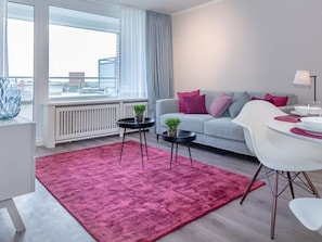 Un salon moderne avec un canapé gris, un tapis rouge, une table à manger avec des chaises blanches et une vue sur la ville grâce à de grandes fenêtres.