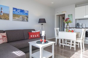 Un salon moderne avec un canapé gris, une petite table blanche avec une bougie, et un coin repas avec une table et des chaises blanches. Deux tableaux encadrés ornent le mur, ainsi qu’une lampe. On aperçoit également un coin cuisine au fond de la pièce.