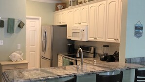 Una cocina con gabinetes blancos, un refrigerador, un microondas, un lavamanos y una encimera de mármol. Hay cuatro taburetes negros y una decoración de pez en la pared.