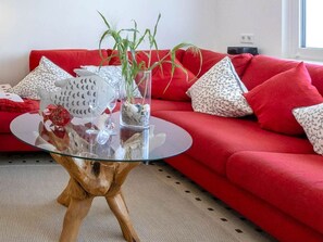 A red sofa with patterned pillows, a glass coffee table with decorative items, and a wooden base.