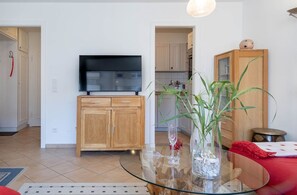 Sala de estar com mesa de centro de vidro, armários de madeira, televisão de tela plana e cozinha ao fundo.
