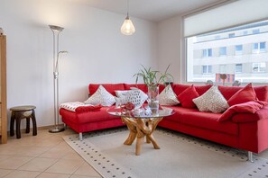 Un canapĂ© rouge avec des coussins Ă motifs, une table basse en verre, un lampadaire et une vue sur un immeuble par la fenĂȘtre.