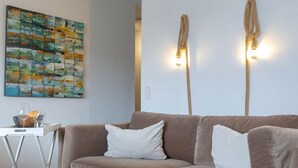 Un salon avec un canapé beige, une table blanche avec un vase, et un tableau abstrait aux couleurs vives accroché au mur.