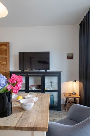 A modern living room with a flat-screen TV mounted on a dark cabinet, a dining table with white plates and a vase of flowers, a grey sofa, and a small wooden side table with a lamp. - Butter with the fishes - beach & city location, modern suite with swimming pool & balcony. (Sylt)