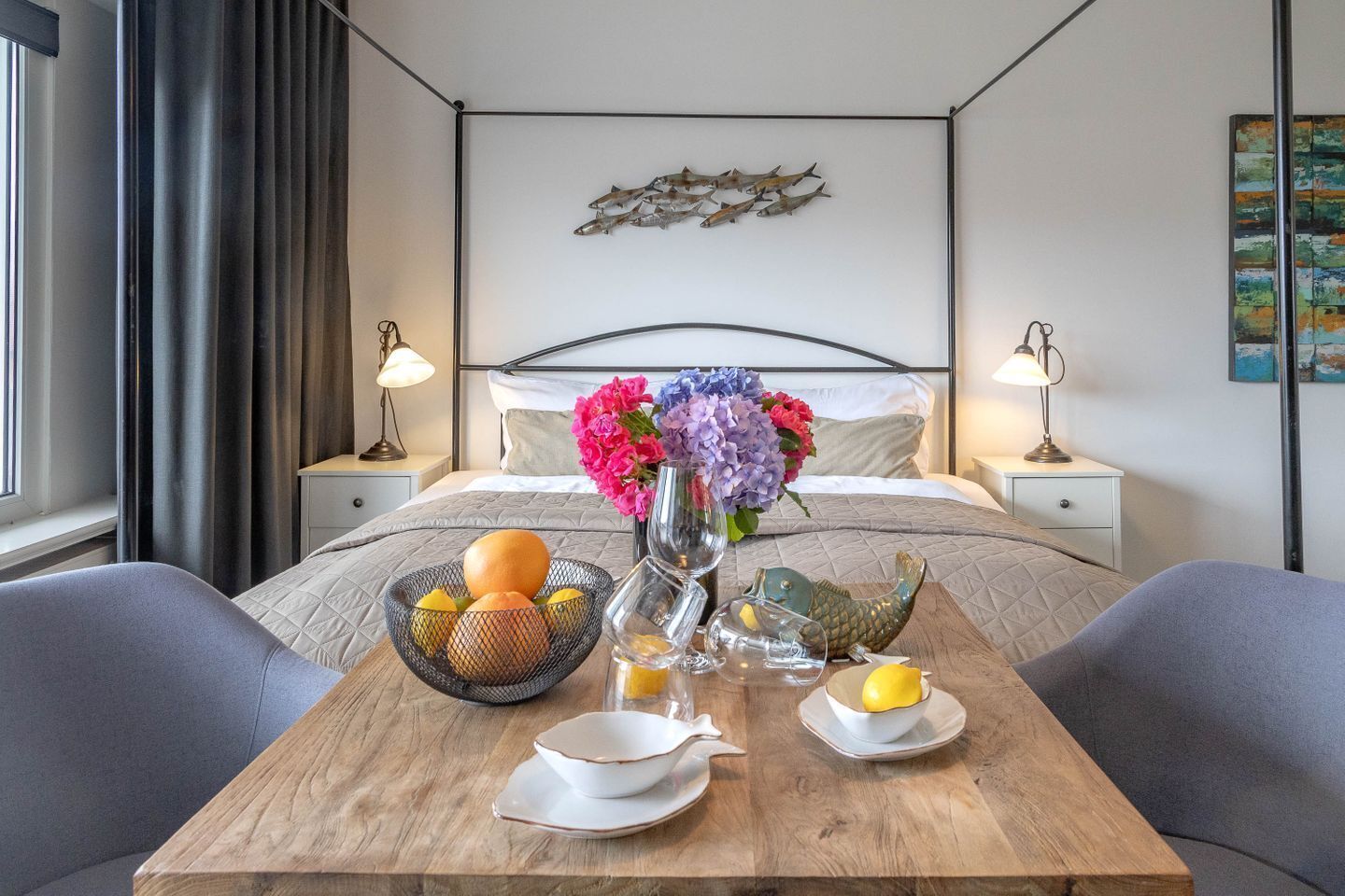 A hotel room with a bed, bedside tables, a wooden table with fruit and a vase, and a wall art piece.