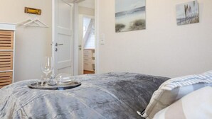 A bedroom with a bed covered in a grey blanket, a tray with glasses and a bottle, and a door leading to another room. - House Senwai - Surfside 2 - Top modern 3 room apartment SURFSIDE 1 in quiet, (Sylt)
