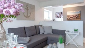 Una sala de estar moderna con un sofá seccional gris, una mesa redonda y una mesita auxiliar blanca con una planta. Hay una obra de arte con forma de ancla enmarcada en la pared y un cuadro sobre el sofá.