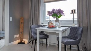 Un comedor moderno con una mesa blanca y dos sillas grises tapizadas. Hay un florero con flores moradas sobre la mesa, y una lámpara es visible a la derecha.
